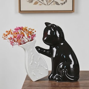 May include: A black ceramic cat figurine with a glossy finish, holding a white vase filled with colorful dried flowers. The vase has a textured surface. Decorative home decor.