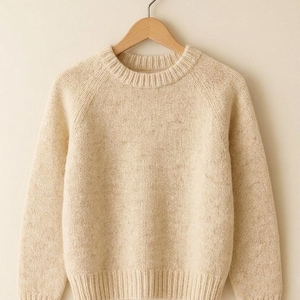 May include: A cream-colored sweater with a crew neck and long sleeves, hanging on a wooden hanger. The knitwear has a ribbed hem and cuffs, and a speckled texture throughout the fabric. The sweater is a neutral tone, suitable for various outfits.