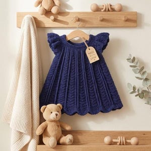 May include: A navy blue knitted baby dress with a scalloped hem and short sleeves hangs on a wooden hanger. A tag reads "Made with Love." A teddy bear and a cream-colored blanket are also visible.