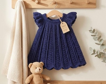 May include: A navy blue knitted baby dress with a scalloped hem and short sleeves hangs on a wooden hanger. A tag reads "Made with Love." A teddy bear and a cream-colored blanket are also visible.