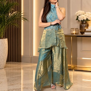 May include: A sleeveless high-neck top in light blue with intricate sequin and bead embellishments, paired with wide-leg trousers in a metallic gold and teal iridescent fabric featuring a layered, flowing silhouette and silver open-toe sandals.