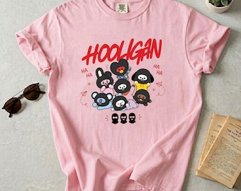 May include: A light pink t-shirt featuring the red text HOOLIGAN above a graphic of several cartoon characters in black masks. The design includes small red text reading HA and various small red line drawings of paper airplanes and stars.