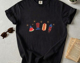 May include: Black t-shirt featuring a graphic of a watermelon slice, a top hat, a hot dog, and a firework. The design is in red, white, and blue, with gold starbursts. The shirt is on a neutral surface.