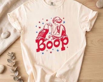 May include: Cream-coloured t-shirt with a red and blue graphic. The design features a woman, an eagle, stars, and the word "Boop" in bold red letters. The shirt has short sleeves and a crew neck.