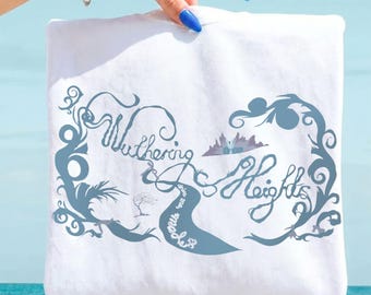 May include: A white t-shirt featuring a blue graphic print with the text Wuthering Heights in a stylised script font. The design includes swirling vine-like patterns, a small tree silhouette, and a mountain range illustration above the text.