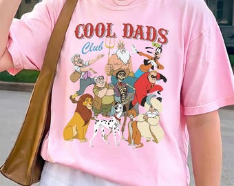 May include: A light pink short-sleeve t-shirt featuring a graphic print with the text COOL DADS Club. The design includes various animated father figures and animal characters from popular films, arranged in a colourful, multi-character collage.