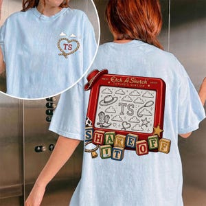 May include: A light blue t-shirt featuring a small rope heart graphic with the letters TS on the front. The back displays a large red Etch A Sketch toy frame with the text Taylor's Version, containing drawings of a hat, boots, and clouds.