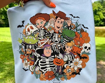 May include: A light blue t-shirt featuring a circular graphic of various cartoon characters wearing skeleton costumes. The design includes pumpkins, spiderwebs, flowers, bats, and a skull, with characters dressed for Halloween in a festive arrangement.