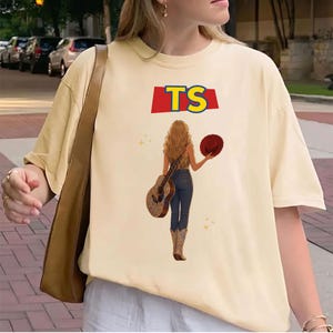May include: A cream-colored short-sleeve t-shirt featuring a graphic of a person with long hair seen from behind, wearing jeans and cowboy boots, holding a guitar and a red hat. Above the figure, the text TS is printed in yellow on a red banner.