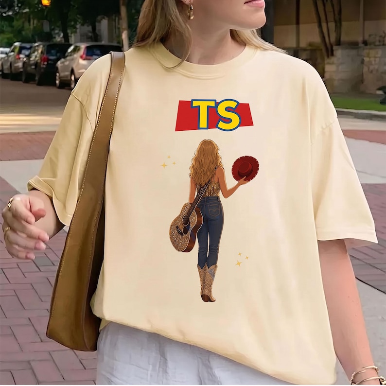 May include: A cream-colored short-sleeve t-shirt featuring a graphic of a person with long hair seen from behind, wearing jeans and cowboy boots, holding a guitar and a red hat. Above the figure, the text TS is printed in yellow on a red banner.