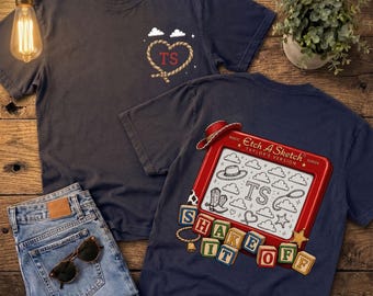 May include: A dark blue t-shirt featuring a rope heart with the letters TS on the front chest. The back displays a red Etch A Sketch toy with the text Taylor's Version, surrounded by cowboy boots, hats, and blocks spelling SHAKE IT OFF.