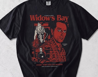 May include: A black short-sleeve t-shirt featuring a red and white graphic print for the Apple TV series Widow's Bay. The design includes a portrait of Matthew Rhys, a clown figure, and the text: Widow's Bay, Every Town Has Its Demons.