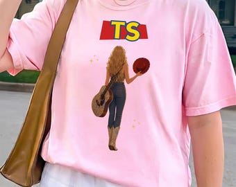 May include: A light pink t-shirt featuring a graphic of a person with long blonde hair seen from behind, wearing dark jeans and cowboy boots. The person holds a brown hat and a guitar. Above the figure, the letters TS appear in yellow on a red background.