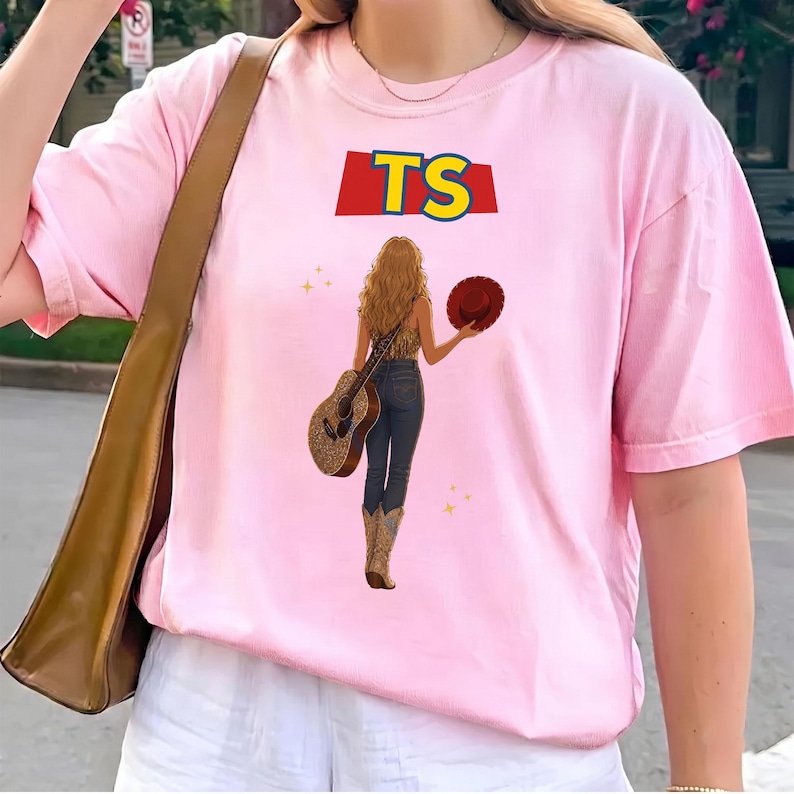 May include: A light pink t-shirt featuring a graphic of a person with long blonde hair seen from behind, wearing dark jeans and cowboy boots. The person holds a brown hat and a guitar. Above the figure, the letters TS appear in yellow on a red background.