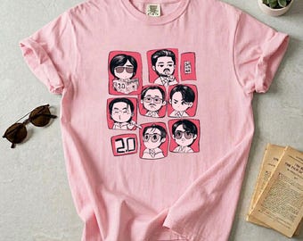 May include: A pale pink t-shirt with a grid of cartoon portraits, each in a pink square. The number "2.0" appears twice. The portraits depict people with different hairstyles and glasses. The word "SAI HANA" is in a small box. Sunglasses are on the left.