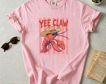 May include: A pale pink t-shirt with a cartoon lobster wearing a cowboy hat and sunglasses, with the text "Yee Claw" in red. The lobster is set against a background of pink and white stripes. The shirt is folded, and sunglasses are visible.