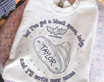 May include: A white crewneck sweatshirt featuring a graphic of a space ranger boot with the name TAYLOR printed on the sole. The design includes the text but I've got a blank space, baby, and I'll write your name, Star Command, and small stars and hearts.