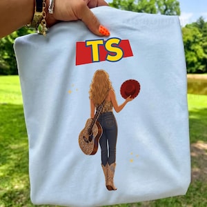 May include: A light blue t-shirt featuring a graphic of a person with long wavy hair seen from the back, wearing a fringed top, dark jeans, and cowboy boots. The person holds a guitar and a red hat. Above, the text TS appears in yellow letters.