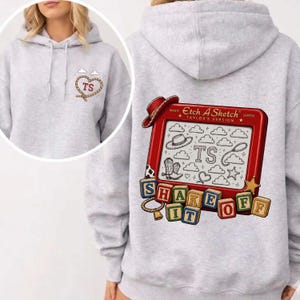 May include: A light gray hooded sweatshirt featuring a small rope heart with the letters TS on the front. The back displays a red Etch A Sketch toy with the text Taylor's Version, containing drawings of clouds, a hat, and boots, with blocks spelling SHAKE IT OFF.
