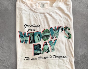 May include: A cream-coloured t-shirt featuring a graphic with the text Greetings from WIDOW'S BAY ...The next Martha's Vineyard! The design includes stylised lettering filled with various illustrations of people and buildings in shades of teal and black.