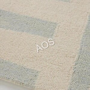 May include: Close-up of a textured rug with a geometric pattern. The rug features alternating sections of light blue and off-white, creating a subtle, modern design. The letters "AOS" are visible on the rug.