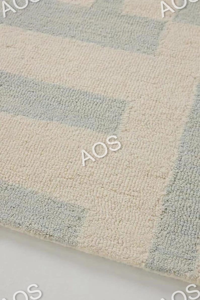 May include: Close-up of a textured rug with a geometric pattern. The rug features alternating sections of light blue and off-white, creating a subtle, modern design. The letters "AOS" are visible on the rug.