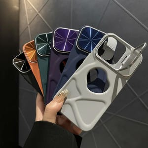 May include: A collection of colorful phone cases in various shades, including gray, orange, teal, purple, and navy blue. Each case features a metallic camera lens protector and a built-in stand for hands-free viewing.