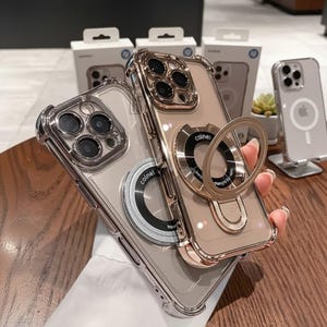 May include: Two clear phone cases with metallic accents and ring holders. One case has a silver frame and ring, the other has a gold frame and ring. The cases are on a wooden surface.