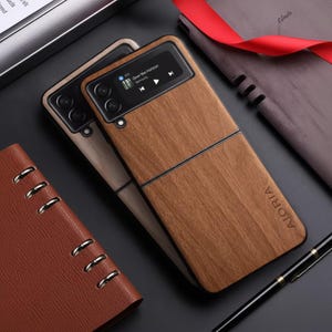 May include: Two wooden-effect phone cases with a black border and the word "AIORIA" printed on the bottom. One case displays a music player interface. A brown leather notebook and a black pen are also visible.