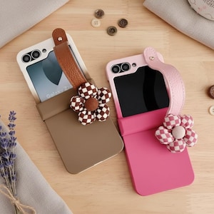 Puede incluir: Dos fundas para teléfono, una marrón y otra rosa, con correas de cuero y adornos florales. La funda marrón tiene una flor a cuadros, y la funda rosa tiene una flor a cuadros rosa y blanca. La pantalla del teléfono muestra el texto "Galaxy Z Flip".