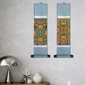 Könnte beinhalten: Zwei vertikale Wandrollen mit aufwendigen, farbenfrohen Designs. Die Rollen haben einen hellblauen Hintergrund mit detaillierten Mustern in Orange, Grün und Gold. Sie haben schwarze Rollen unten und kleine silberne Haken oben.