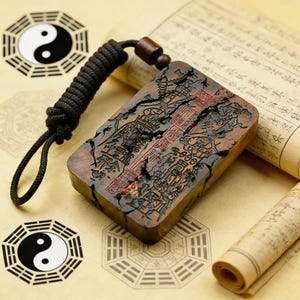 May include: A rectangular wooden pendant with intricate carvings and a black cord. The pendant is a reddish-brown color with detailed relief carvings. A black cord is attached to the pendant, and two Yin Yang symbols are visible in the background.