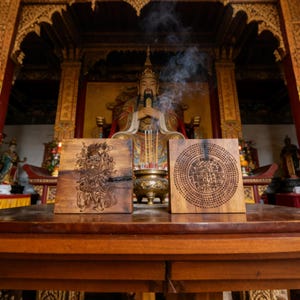 Puede incluir: Dos placas cuadradas de madera con diseños intrincados se exhiben sobre una mesa de madera. Una placa presenta una ilustración detallada de una criatura mítica, mientras que la otra muestra un patrón circular de símbolos. Humo de incienso en el fondo.