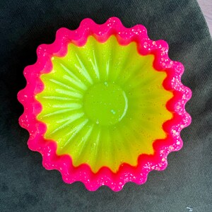 May include: A vibrant, decorative bowl with a scalloped edge. The bowl features a bright neon pink rim with glitter accents, surrounding a yellow and green interior. The bowl has a flower-like design and is set against a dark background.
