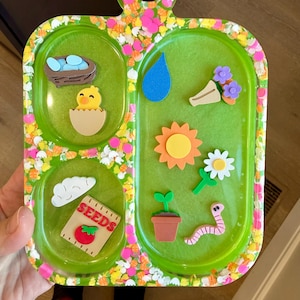 May include: A green tray with a handle, decorated with colorful floral confetti. The tray features spring-themed cutouts, including a chick hatching from an egg, a nest with blue eggs, a water droplet, a sun, and a packet of seeds.