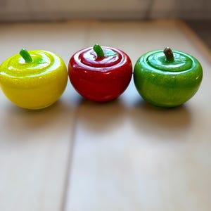 May include: Three round, apple-shaped containers with lids, arranged in a row. The left container is bright yellow, the center is vibrant red, and the right is lime green. Each features a small, sculpted stem on top and a smooth, glossy finish.