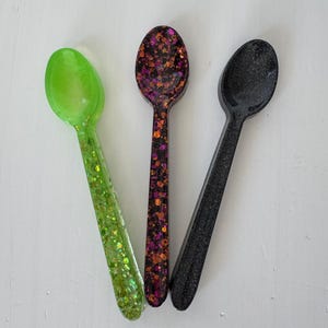 May include: Three decorative spoons in a close-up shot. One is lime green with glitter, the second is dark purple with orange and pink glitter, and the third is black with glitter. The spoons are arranged on a white surface.