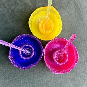 May include: Three colorful resin spoon rests. Each rest is a different color: yellow, blue, and pink. Each spoon rest has a matching spoon. The edges of the spoon rests are decorated with glitter.