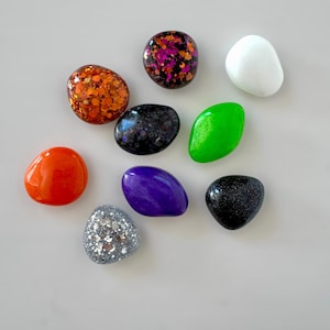 May include: A collection of colorful, smooth, pebble-shaped decorative stones. The stones vary in color, including orange, purple, green, white, and black, some with glitter accents. These decorative stones are ideal for crafts and home decor.