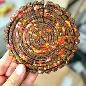 May include: A circular coaster with a textured brown surface mimicking the appearance of a tree stump cross-section. The coaster features concentric rings and is embedded with scattered, reflective glitter in shades of orange, pink, and yellow.