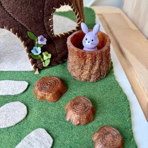 May include: A felt treehouse with a small purple bunny figurine inside a tree stump. The scene includes felt tree stumps and white oval shapes on a green felt base, creating a whimsical forest setting.