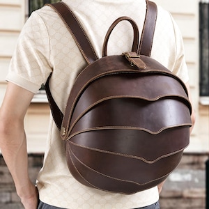 May include: A brown leather backpack with a unique layered design. The backpack has adjustable shoulder straps and a top handle. The bag is secured with a buckle closure. The stitching is visible, adding to the handcrafted aesthetic. The backpack is worn on the back.