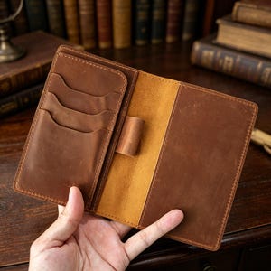 Puede incluir: Diario de cuero marrón abierto con múltiples ranuras para tarjetas y un portalápices. El diario está abierto, revelando un interior beige y un exterior de cuero envejecido. Las costuras son visibles alrededor de los bordes. El diario se sostiene en una mano.