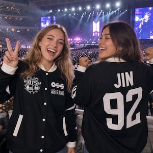 May include: Two people wearing black and white jackets at a concert. One jacket has the text "BTS" and "ARIRANG" printed on it. The other jacket has the name "JIN" and the number "92" printed on it. The background shows a large crowd and stage.