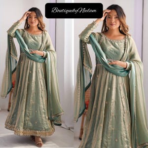 May include: Sage green formal dress with long sleeves and a flowing skirt. The dress features delicate embroidery and a matching teal scarf. The text "BoutiquebyNeelam" is displayed in a black box at the top. The dress has a gold trim at the bottom.