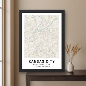 May include: A framed map print of Kansas City, Missouri, USA, featuring a light beige background with a winding blue river and black street grid lines. The text at the bottom reads KANSAS CITY, MISSOURI, USA, 39.09518 N, -94.59055 E.