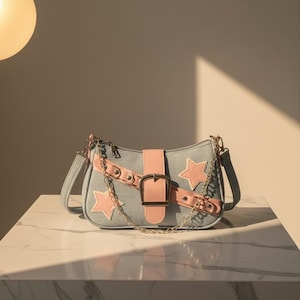 Peut inclure: Sac à bandoulière en jean bleu clair avec une sangle et une boucle en cuir rose. Le sac comporte des patchs en forme d'étoile roses et une bandoulière en chaîne dorée. Le sac est posé sur une surface en marbre blanc.