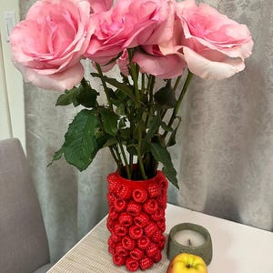 Può includere: Un bouquet di rose rosa in un vaso rosso con un design a lampone. Il vaso è su un tavolo bianco accanto a una candela e una mela gialla. Le rose hanno grandi petali morbidi.