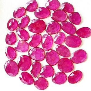 Puede incluir: Una colección de numerosas piedras preciosas de color rojo rosado, con forma ovalada y talladas en facetas, dispuestas en un grupo suelto. Cada piedra presenta una superficie pulida con múltiples facetas geométricas que reflejan la luz, mostrando un tono magenta vibrante.