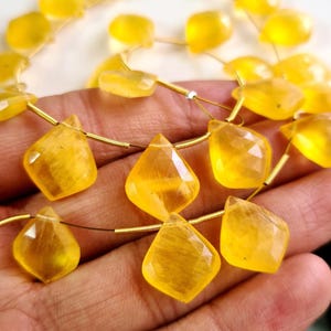 May include: A strand of faceted, translucent yellow gemstone beads shaped like diamonds. The beads are strung on a thin, metallic wire, showcasing a bright, sunny hue and a polished, reflective surface that catches the light across each cut facet.